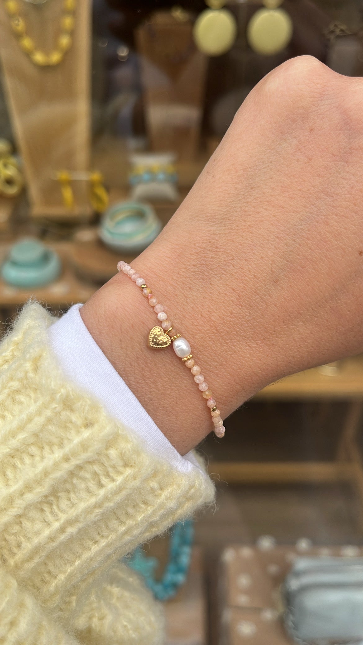 Poignet de femme portant un bracelet avec des petites perles rose, une perle blanche et un petit coeur en acier inoxydable doré