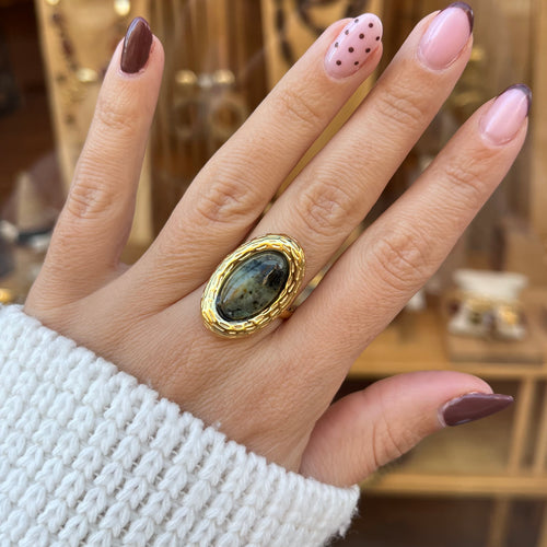 Main de femme portant Une bague de forme ovale en acier inoxydable doré, avec une pierre verte au centre 