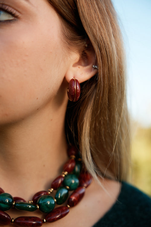 Visage d’une femme portant des petites boucles d’oreilles en résine bordeaux, ainsi que 3 colliers en résine