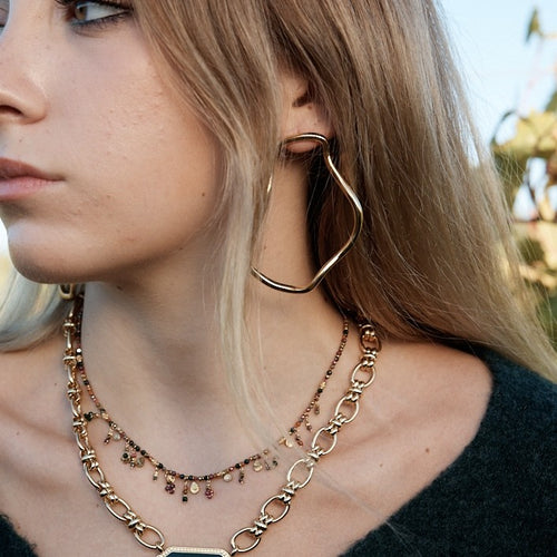 Visage d une femme portant une boucle d oreille dorée en acier inoxydable, ainsi que 2 colliers superposés 